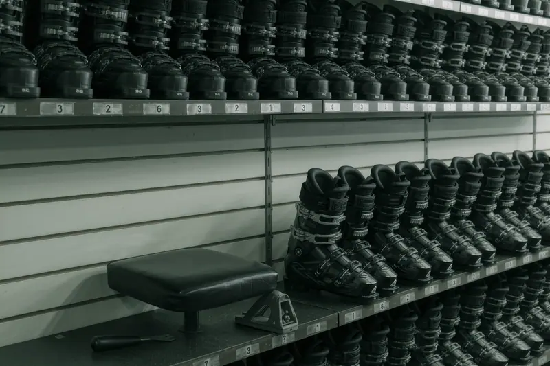 Ski boots on a heated rack beside a fitting stool, with a shell punch and small hand tools laid out on the bench behind.