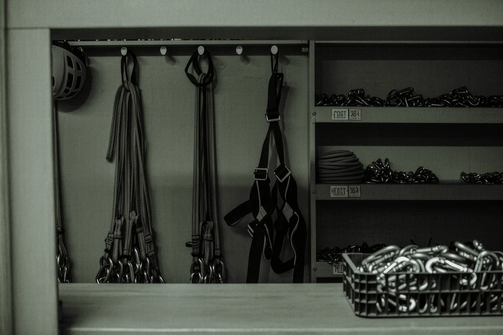 Climbing harnesses, via ferrata lanyard sets and helmets hanging on a pegged rental wall, an open crate of carabiners standing on the counter in the foreground.