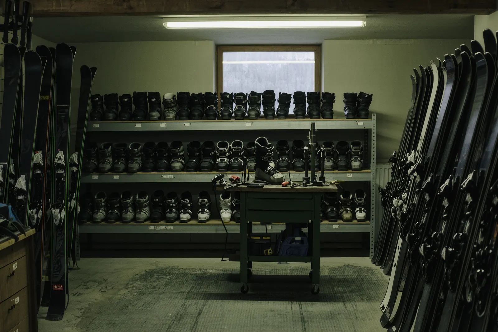 The workshop of a ski rental shop, skis in numbered wall racks and boots lined up on a steel shelf beside a fitter's bench.