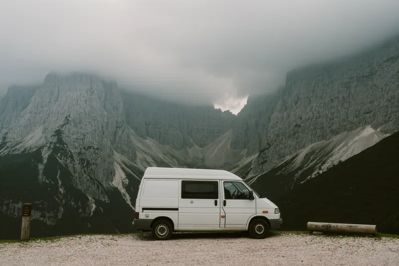 A compact campervan parked on a gravel pull-in below grey limestone peaks, the road empty beyond it.