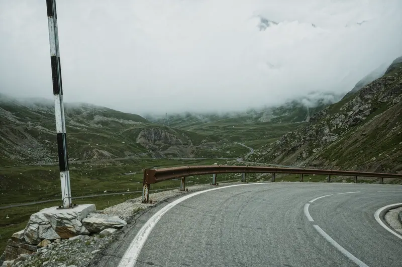 An empty alpine road with a crash barrier on the outer edge and a rock cutting on the inside, under flat grey cloud.