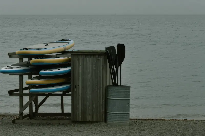 Paddleboards stacked on a rack beside a lake changing hut, with paddles standing upright in a barrel.