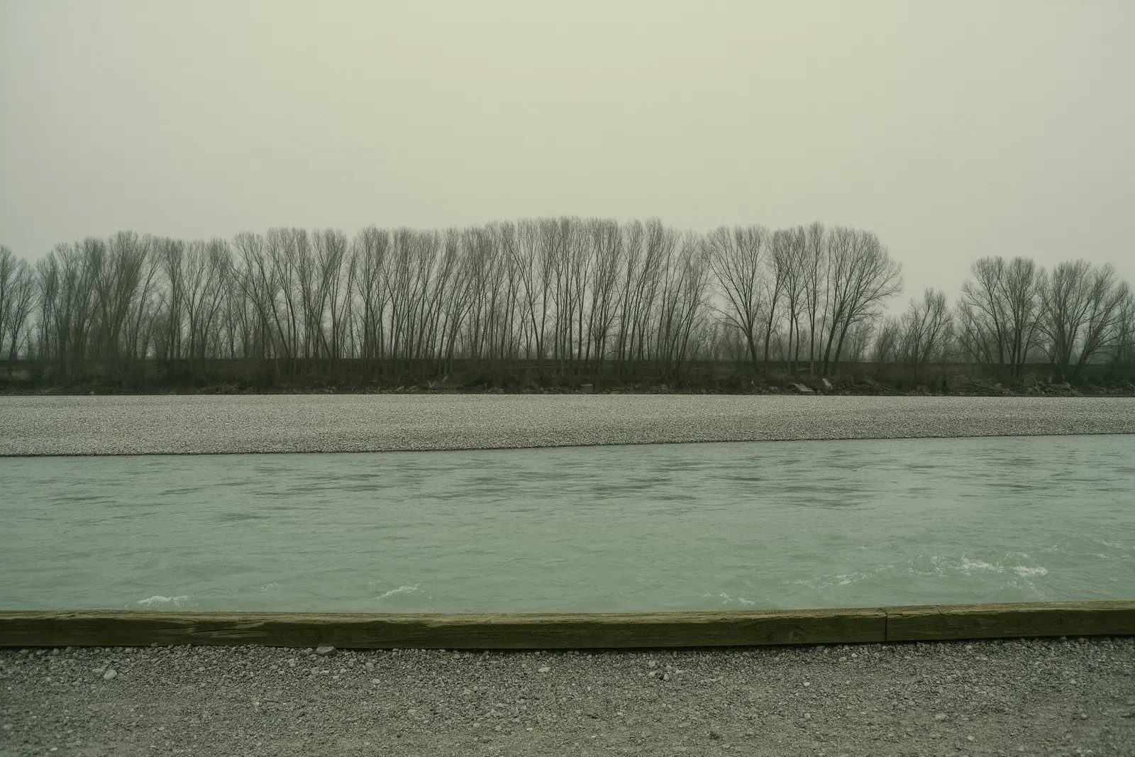 A gravel riverside cycle path beside a fast pale-green river, loose shingle and a weathered wooden edging beam in the foreground, bare trees and a shingle bank behind.